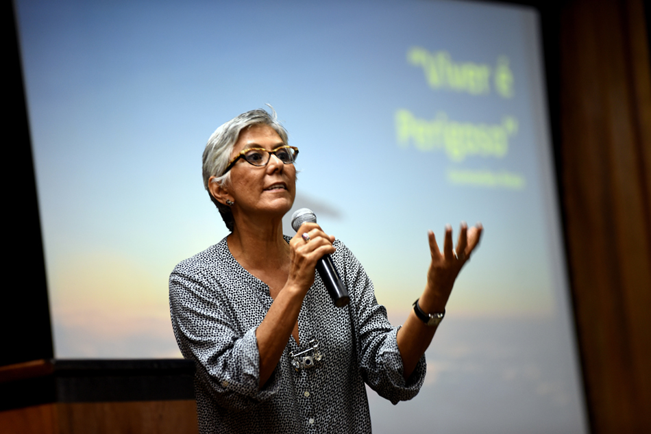 A jornalista e consultora Olga Curado fala, de pé, ao microfone, onde se lê, atrás dela, em uma tela, a frase "viver é perigoso", durante o Simpósio "Comunicação: Universidade e Sociedade", realizado dia 7 de dezembro, no Centro de Convenções da Unicamp