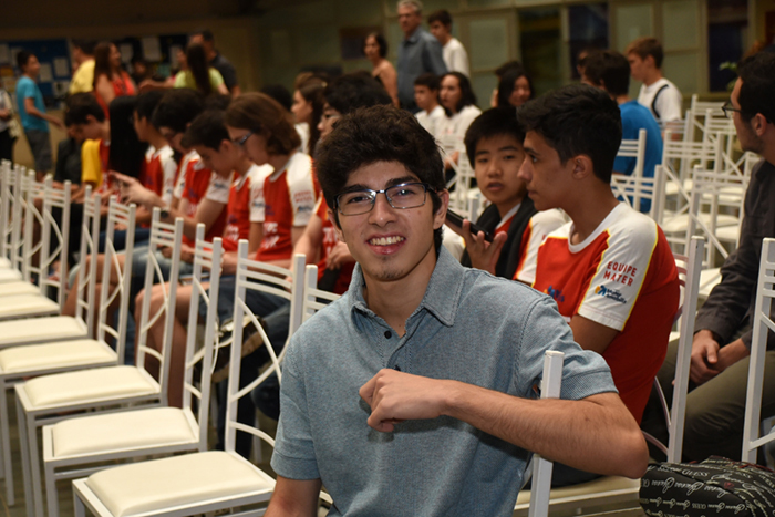 Fileira de cadeiras na diagonal com um jovem, medalhista de ouro na 32ª Olimpíada de Matemática da Unicamp, sentado à direita, com público ao fundo