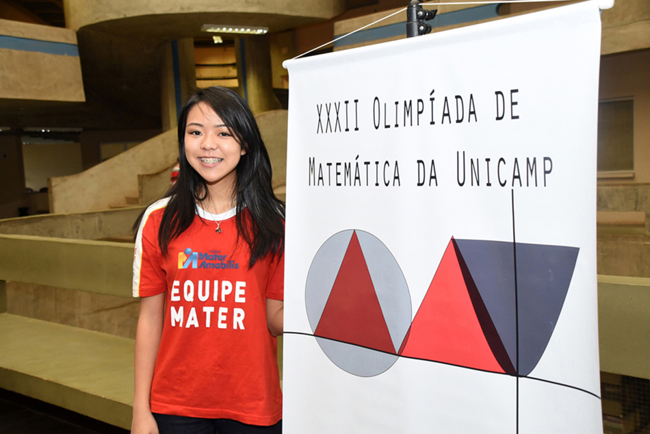 Uma jovem, à esquerda, com banner de divulgação sobre a 32ª Olimpíada de matemática da Unicamp ao seu lado, à direita
