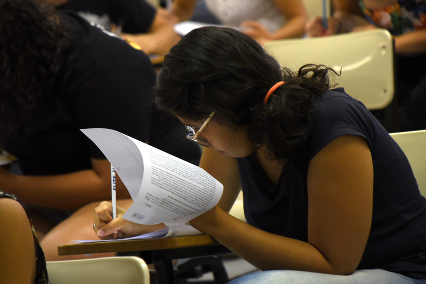 Em sala de aula, candidata, em primeiro plano, realiza prova utilizando carteira escolar, com outros candidatos ao fundo