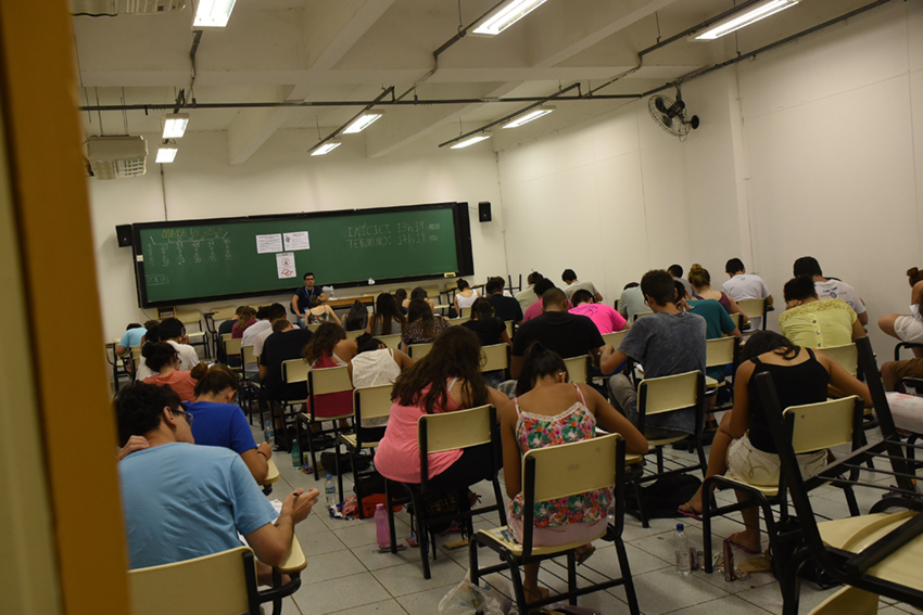 Sala de aula, em perspectiva, com cerca de 50 pessoas, de costas, acomodadas em carteiras escolares realizando provas do vestibular, tendo, à frete, quadro negro e fiscal de provas