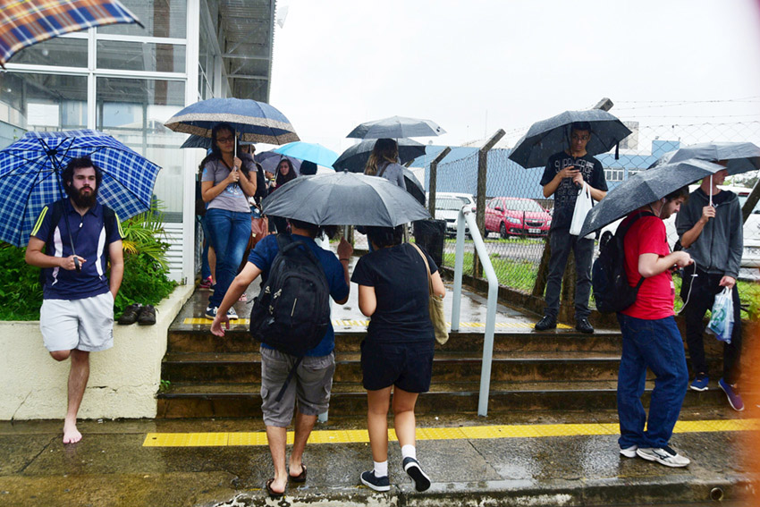 Várias pessoas usando guarda-chuva nas proximidades de uma escada de três degraus que dá acesso ao local das provas
