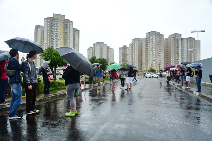 Várias pessoas usando guarda-chuva ao longo de uma rua, em perspectiva, com altos prédios ao fundo