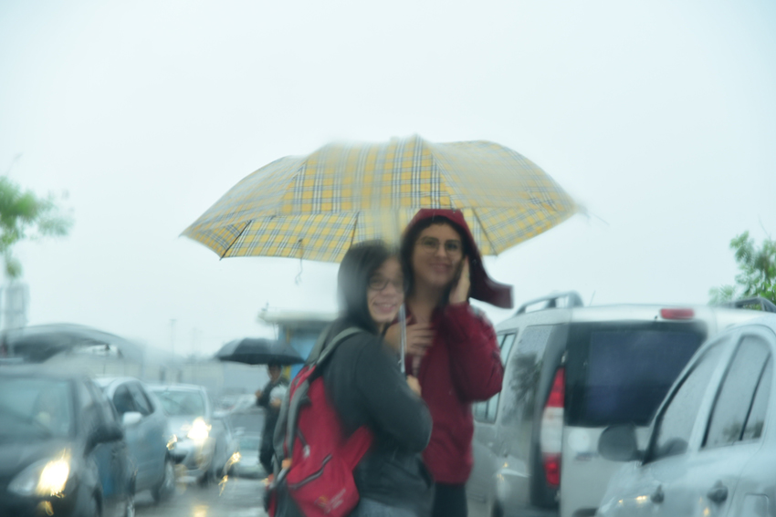 Duas mulheres paradas sob o mesmo guarda-chuva com carros circulando à esquerda e à direita em sentidos opostos
