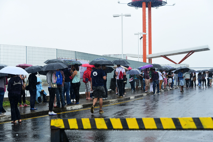 Várias pessoas usando guarda-chuva, paradas na calçada, em perspectiva, com uma grade metálica à esquerda e um grande portão de entrada à frete