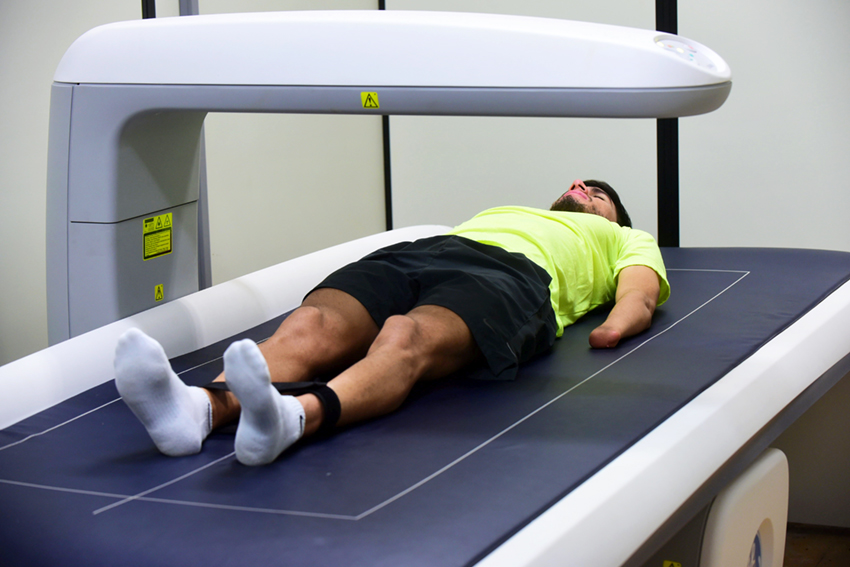 Homem (atleta), em perspectiva, deitado em equipamento com parte plana, semelhante a uma esteira, com braços alinhados às pernas, em sala com divisórias de madeira