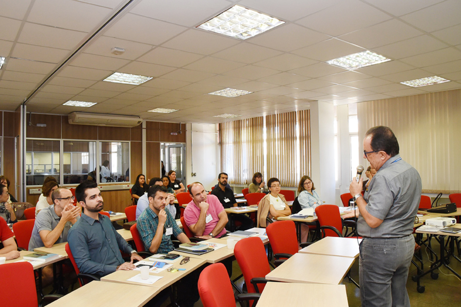Em perspectiva, sala de aula com cerca de 30 pessoas, entre homens e mulheres, sentados em cadeiras e tendo, à frente, mesas de madeira dispostas, uma junto à outra, em semi-círculo. Ao fundo, no final da sala,, há uma grande janela de vidro, e na lateral, à direita, outra grande janela de vidro dotada de percianas verticais, sendo que à frente do grupo há um homem, em pé, se comunicando com o grupo