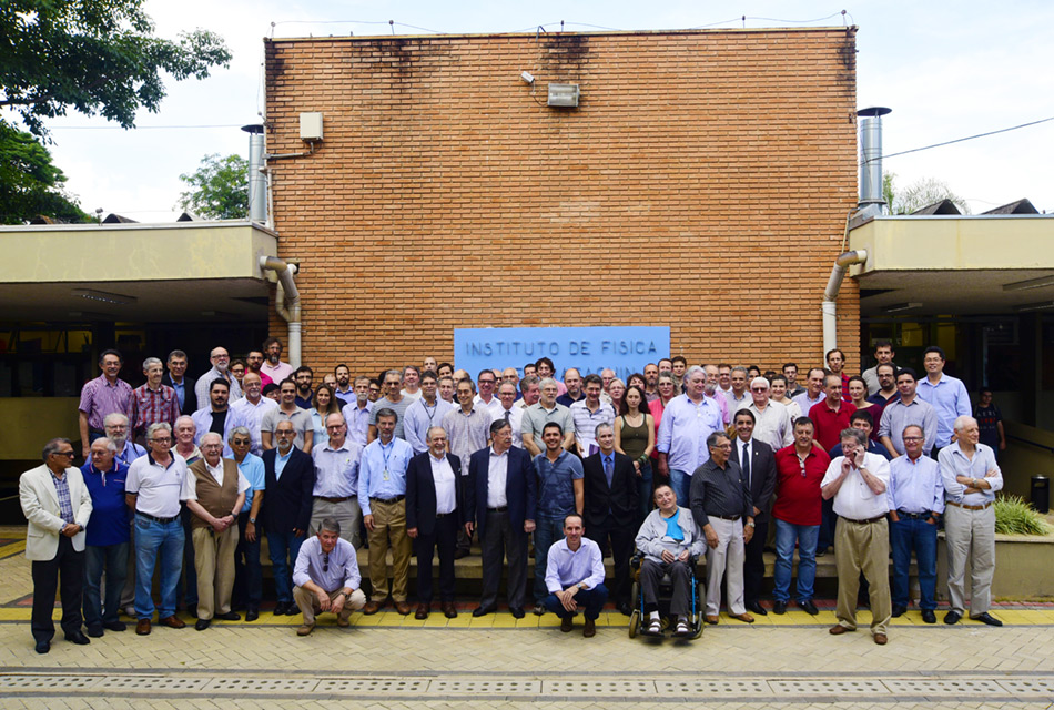 Imagem frontal, à distância, grupo de cerca de 100 pessoas, sendo 90 por cento delas homens e 10 por cento mulheres, em pé, exceto 2 que estão agachados e um cadeirante, à frente do grupo, posicionados um ao lado do outro, formando três fileiras. Eles estão à frente de uma edificação, sendo que atrás deles há uma grande parede de tijolinhos, formando a fachada do prédio, com cerca de sete metros de altura por sete de largura, onde há no centro uma placa retangular em tom azul, onde se lê instituto de física
