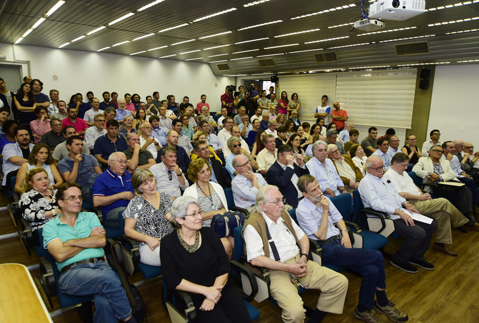 Em perspectiva, em um auditório, cerca de oitenta pessoas entre homens e mulheres, sentados em cadeiras, sendo que ao fundo e em uma lateral há pessoa agrupadas em pé