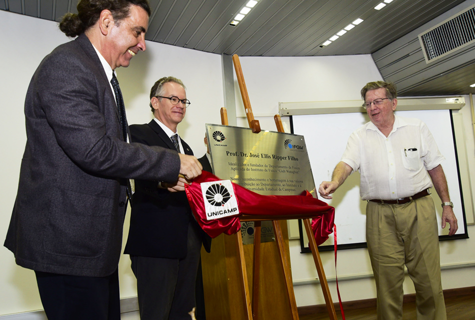 Em perspectiva, três homens, em palco de um auditório, sendo que dois deles, à esquerda, trajam terno e gravata, e o outro, à direita, camisa de botões de mangas curtas e calça social, desceram placa de metal retangular, que está à frente deles, apoiada sobre cavalete de madeira, onde se lê homenagem ao professor doutor José Ellis Ripper Filho