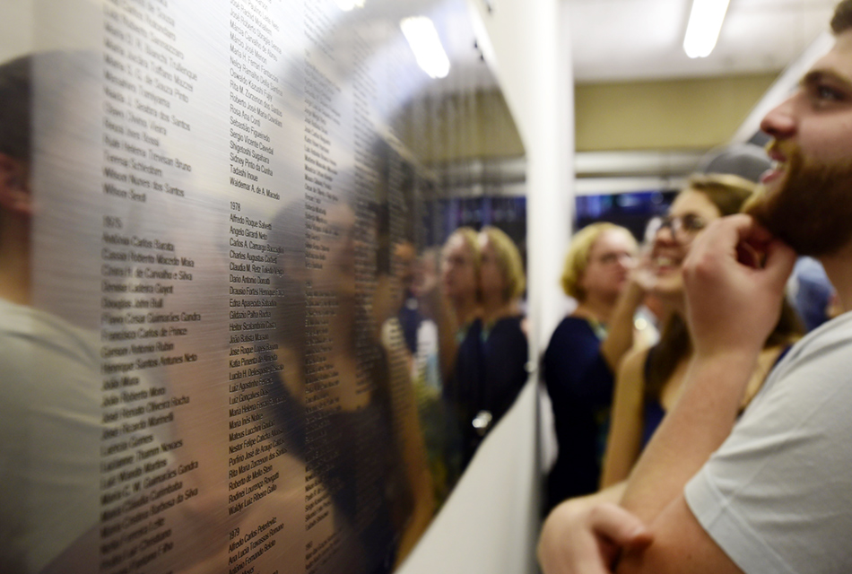 Em perspectiva, um extenso painel metálico fixado em parede, à esquerda, com a gravação dos nomes de ex-alunos do instituto, alinhados em colunas e abertura com o ano das turmas, que são observadas por várias pessoas que estão à direita, com expressão de alegria