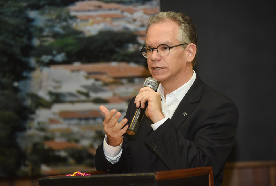 Em um púlpito de madeira de um amplo auditório, homem, em pé, fala ao microfone sem fio, segurando-o com a mão esquerda. Ele traja roupa social e usa óculos, sendo que às costas há um grande painel com uma foto aérea de área da Unicamp, à esquerda