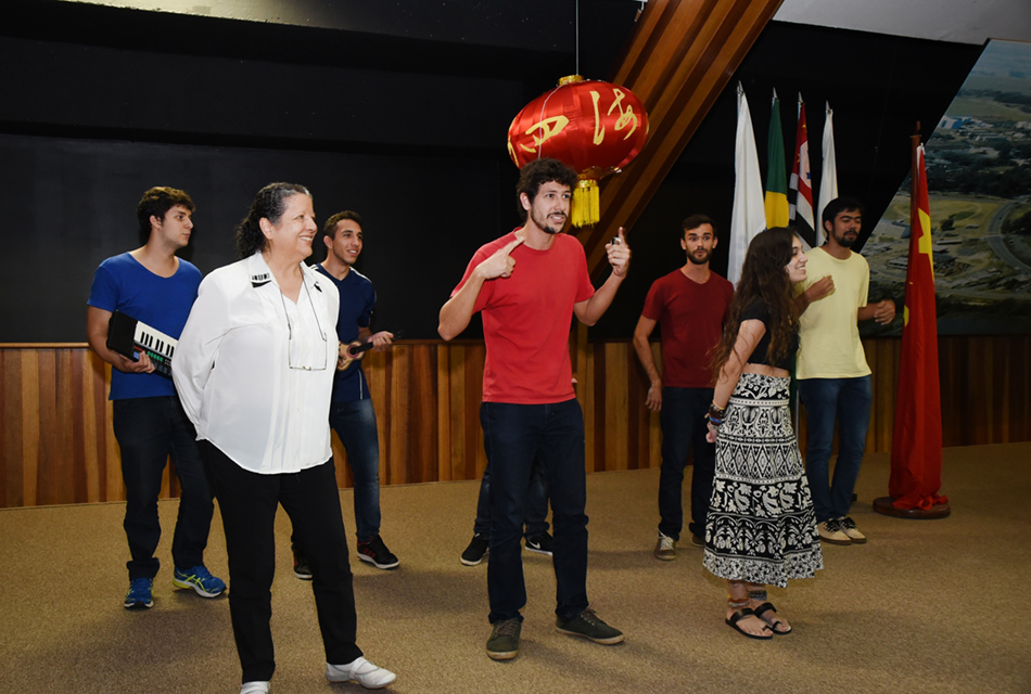 Grupo de oito pessoas, entre homens e mulheres, em pé, em um amplo palco acarpetado de um auditório, fazem apresentação de música chinesa, sendo que dois deles tocam instrumentos musicais. Acima deles há um balão vermelho em estilo chinês, e ao fundo, à direita, há, em destaque, uma bandeira chinesa, e também as bandeiras da Unicamp, Campinas, Estado de São Paulo e do Brasil, todas fixadas em mastros