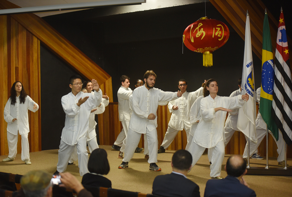 Grupo de 12 pessoas, entre homens e mulheres, em pé, em um amplo palco de um auditório, fazem apresentação de Taijiquan (Tai Chi Chuan), todos em conjunto, com movimento inclinando o corpo para frente, colocando um pé para frente e outro para trás, e os braços levantados à frente, como se segurassem uma grande bola, com delicadeza. Eles vestem roupa branca apropriada à atividade. Acima, balão vermelho em estilo chinês, e à direita, as bandeiras de Campinas, Estado de São Paulo e do Brasil fixadas em mastros