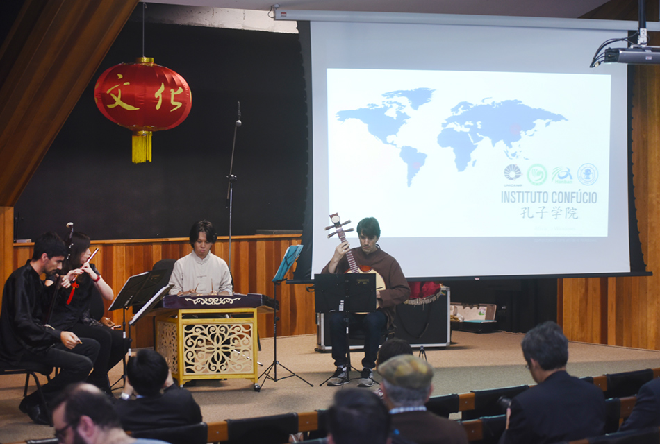 Em um amplo palco de um auditório, quatro pessoas, sentadas em cadeiras, formando um semi-círculo, de frente para a plateia, fazem apresentação musical vestindo roupas típicas chinesas e tocando instrumentos típicos chineses, de corda e sopro. Acima deles, há um balão vermelho em estilo chinês, e ao fundo, à direita, uma grande tela de projeção de data show que exibe o mapa mundi, logotipos de instituições e também se lê Instituto Confúcio, escrito em português e chinês