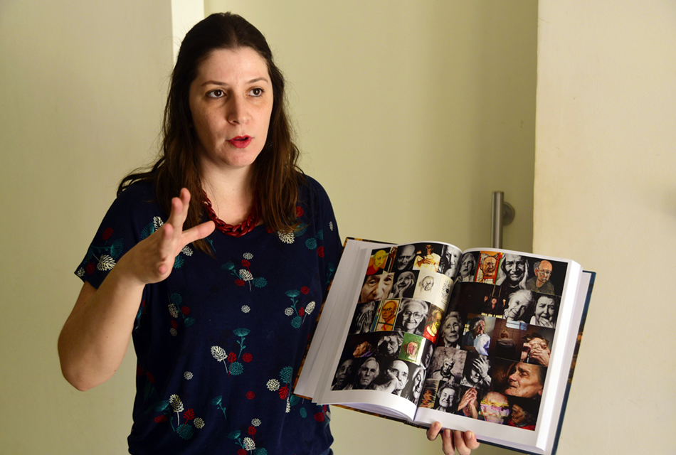 Em uma sala, mulher em pé, segurando com a mão esquerda um livro aberto, de forma a mostrar duas páginas onde aparecem várias fotos pequenas e coloridas de rostos de pessoas, sendo que ela gesticula com o braço direito, erguendo-o próximo ao corpo na altura do pescoço, com a mão aberta. Ela veste uma blusa em tom preto  com pequenos detalhes coloridos e usa um colar vermelho de pano
