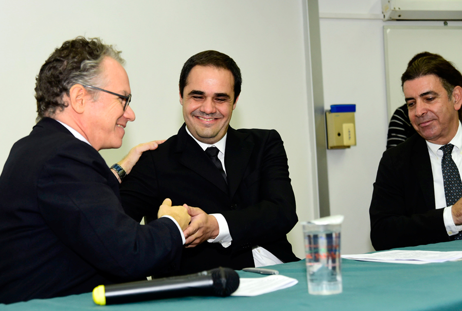 Em auditório, três homens sentados em cadeira, um ao lado do outro, tendo, à frente, uma extensa mesa de madeira recoberta por tecido em tom verde sobre a qual há um microfone sem fio, um copo plástico com água e folhas de papel sulfite. Os dois homens à esquerda se cumprimentam sorrindo e com aperto de mãos, observados pelo outro homem, à direita. Um deles usa óculos e todos trajam terno e gravata em tom preto