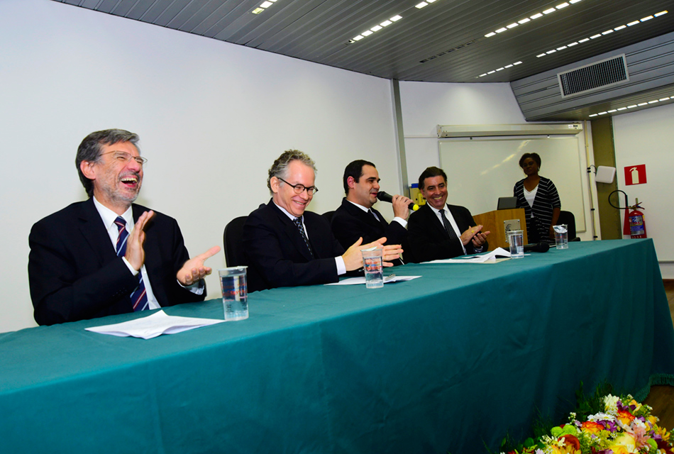 Em auditório, quatro homens sentados em cadeira, um ao lado do outro, tendo, à frente, uma extensa mesa de madeira recoberta por tecido em tom verde sobre a qual há copos plásticos com água e folhas de papel sulfite. Um dos homens ao centro fala em microfone, sendo que todos trajam terno e gravata em tom preto