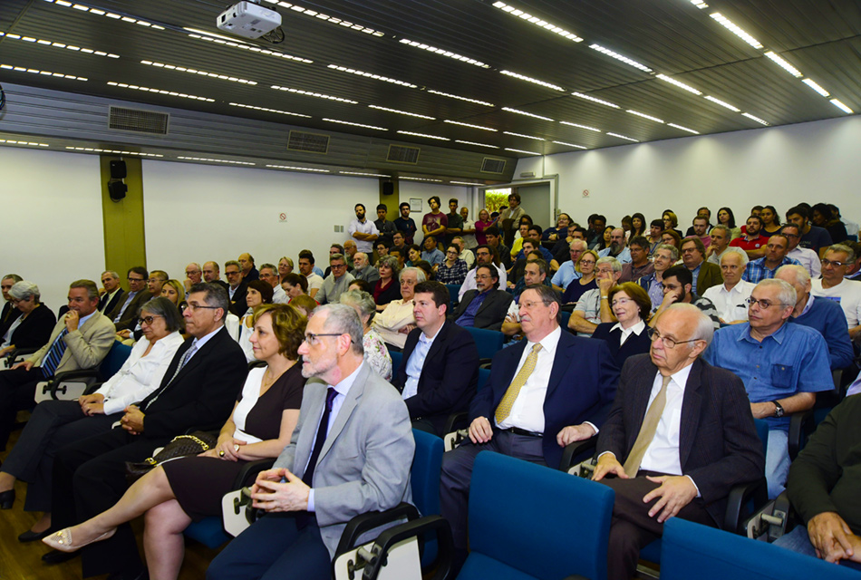 Em amplo auditório, em perspectiva, cerca de 100 pessoas, entre homens e mulheres, sentados em cadeiras alinhadas em fileiras horizontais e verticais, em desnível, sendo que a maioria veste roupa social