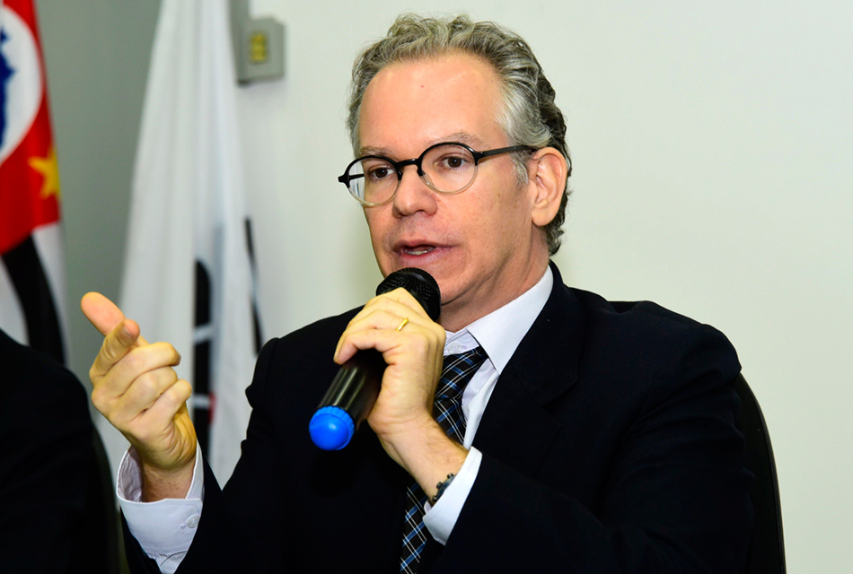 Em auditório, homem sentado fala em microfone sem fio segurando-o com a mão esquerda, enquanto gesticula com a mão direita, com que apontando para algo. Ele usa óculos e traja terno e gravata em tom preto, tendo às costa quatro bandeiras de pano hasteadas em pequenos mastros
