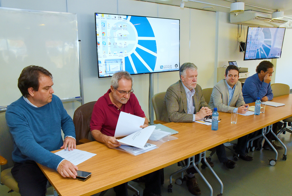 Em pequeno auditório, cinco homens sentados em cadeiras, um ao lado do outro, tendo à frente uma extensa mesa de madeira retangular, sobre a qual apoiam os braços. Sobre a mesa, há vários blocos de folhas de sulfite impressa, e Às costas, há dois grandes monitores de tv led fixados na parede onde aparecem os logos da Unicamp e do Instituto de Economia