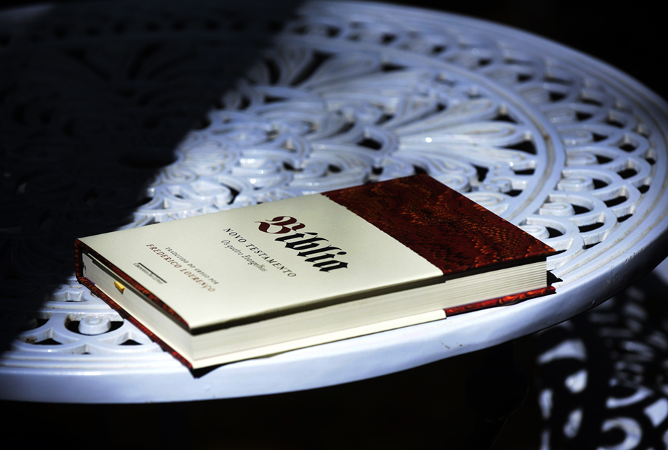 Em ambiente de penumbra, livro impresso, sobre uma mesa redonda de ferro, vazada, onde se lê na capa Bíblia - Novo Testamento Quatro Evangelhos