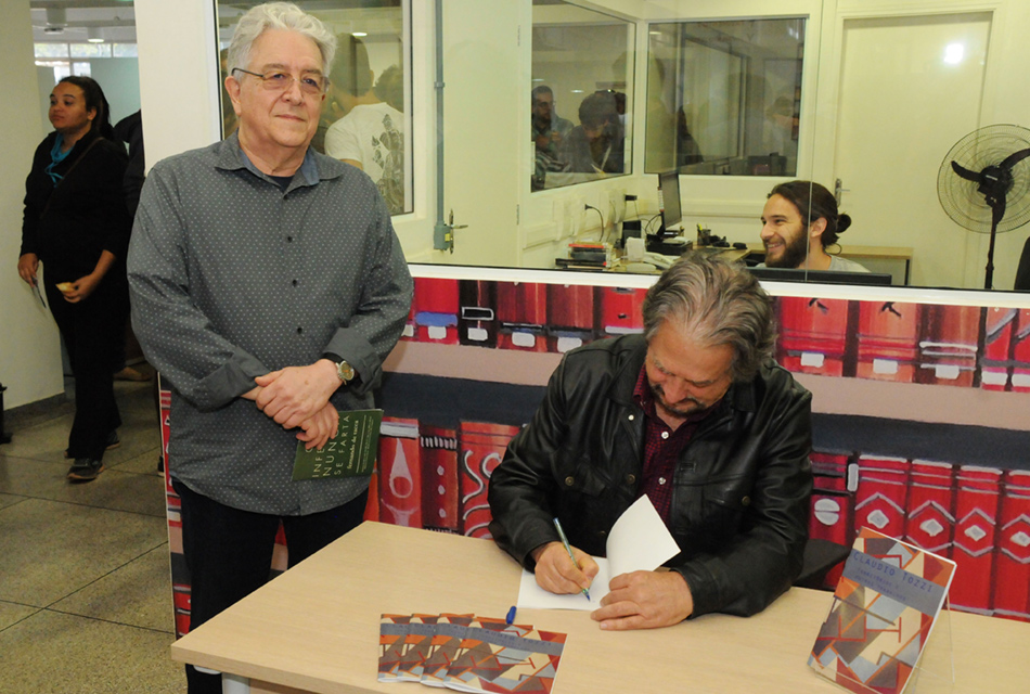 Dois homens, um ao lado do outro, sendo que o da esquerda está em pé, com as mãos unidas na altura do ventre; e o outro sentado em cadeira, com uma mesa de madeira retangular à frente, onde autografa um livro aberto, tendo alguns exemplares sobre a mesa