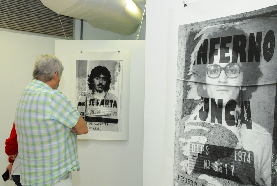 Em sala de exposição, pessoas em pé observam trabalhos artísticos afixados em paredes, sendo que o material, cartaz impresso com cerca de cinquenta centímetros de largura por setenta de altura, traz fotos em preto e branco de pessoas, estilizado, com tom verde no entorno, onde se lê a frase o inferno nunca se farta