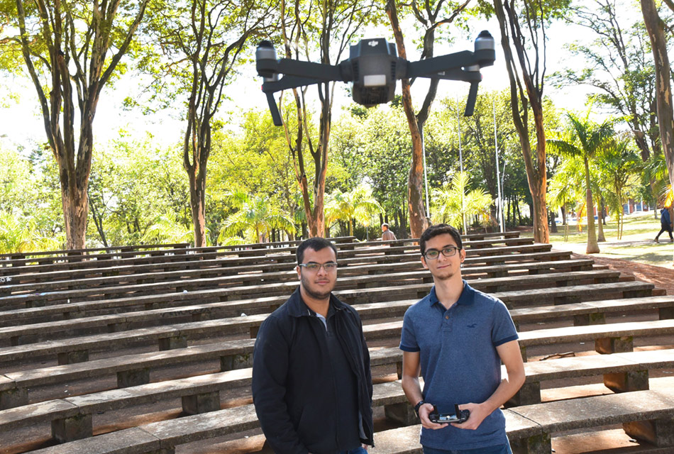 Em área externa, dois jovens, em pé, um ao lado do outro, sendo que o da direita opera um controle remoto com as duas mãos, tendo acima, voando ao alto, o drone, equipamento com aproximadamente setenta centímetros de diâmetro, com hélices nas pontas de quatro eixos que se unem a uma peça central retangular onde estão os sensores e também uma micro câmara de foto e filmagem à frente. Às costas deles, há uma arquibancada baixa de concreto, construída em formato circular e com altas árvores no entorno