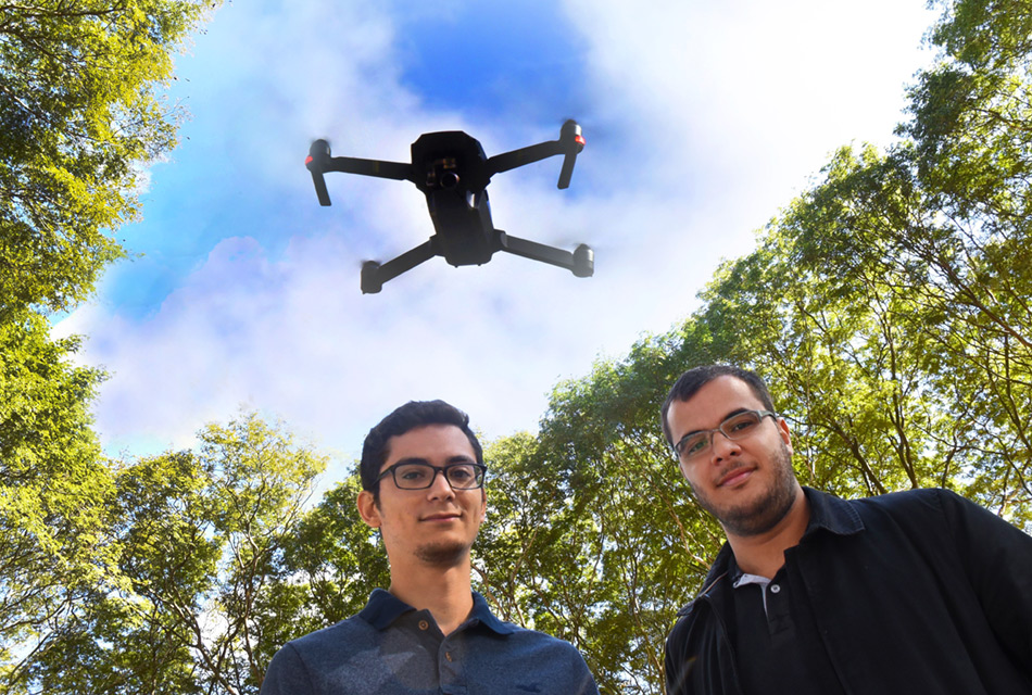 Em área externa, dois jovens, em pé, um ao lado do outro, tendo acima, voando ao alto, o drone, equipamento com aproximadamente setenta centímetros de diâmetro, com hélices nas pontas de quatro eixos que se unem a uma peça central retangular onde estão os sensores e também uma micro câmara de foto e filmagem à frente. No entorno deles há altas árvores