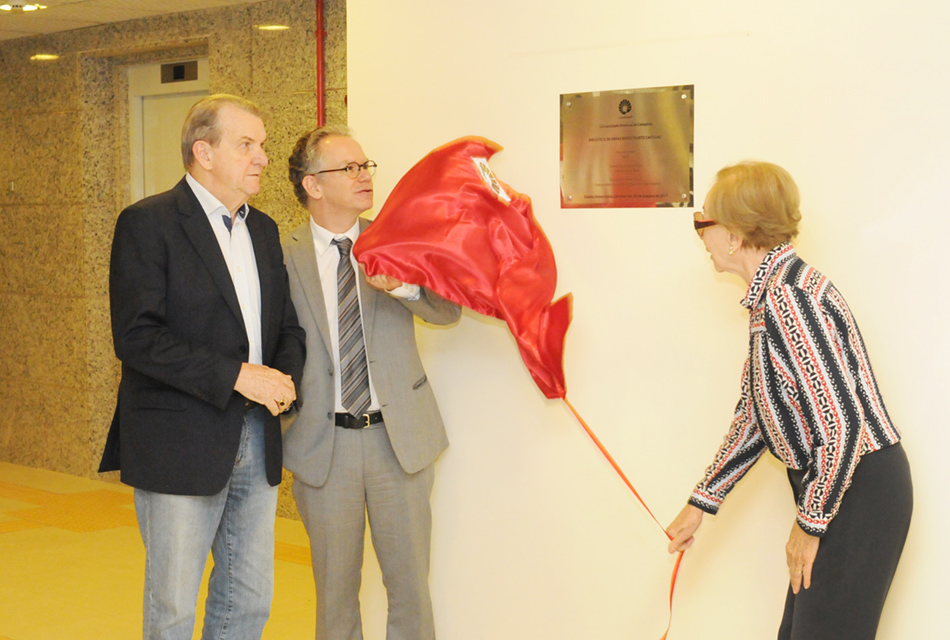A galeria traz a cerimônia de nomeação da Biblioteca Fausto Castilho, na Unicamp. Para acessá-la, tecle enter. Audiodescrição: Em sala, mulher, à direita, descerra placa metálica prateada, com cerca de quarenta centímetros de largura por cinquenta de altura, que encontra-se afixada na parede, puxando para baixo, com a mão direita, uma corda presa a um pano vermelho. Ela é auxiliada por um homem que está à sua frente, à esquerda. Há um outro homem que participa do descerramento, à esquerda. Imagem 1 de 4.