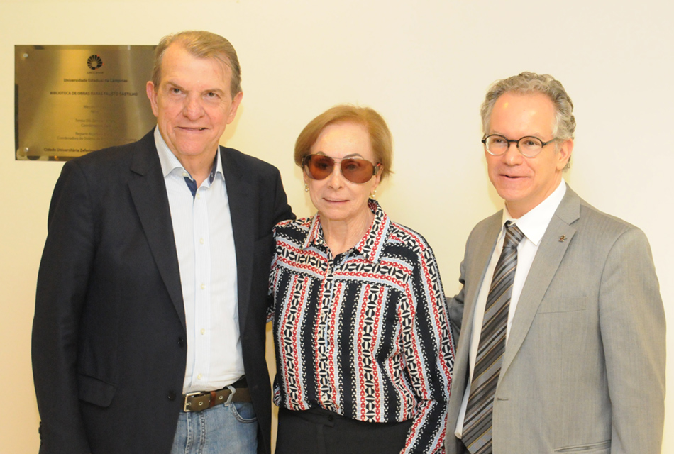 Em imagem frontal, dois homens e uma mulher, ao centro, em pé, um ao lado do outro, sorrindo. Eles vestem blazer sobre camisa social, e o homem à direita, gravata, enquanto a mulher usa óculos escuros e veste camisa de botões de mangas compridas com listras verticais pretas e rosas. No alto, à esquerda, há uma placa metálica prateada afixada na parede, com cerca de cinquenta centímetros de largura por quarenta de altura. Imagem 2 de 4.