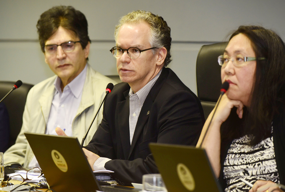 A galeria mostra reunião do Conselho Universitário da Unicamp. Para acessá-la, tecle enter. Audiodescrição: Em auditório, em perspectiva, três pessoas sentadas em cadeira, uma ao lado da outra, sendo dois homens e uma mulher, à esquerda. À frente deles, há uma extensa mesa, sobre a qual há notebooks e microfones de mesa à frente de cada um, sendo que o homem ao centro fala em um deles. Todos usam óculos, os homens vestem blazer sobre camisa social, e a mulher blusa estampada em preto e branco. Imagem 1 de 6