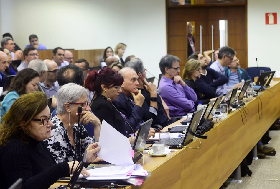 Em amplo auditório, em perspectiva, cerca de trinta pessoas sentadas em cadeiras, uma ao lado da outros, formando cinco fileiras, sendo que, à frente deles, há extensas mesas retangulares de madeira sobre a qual há notebooks abertos, microfones de mesa, folhas de sulfite entre outros objetos. À direita na imagem, ao lado deles, há uma grande porta balcão de madeira com duas lâminas e janelas verticais de vidro transparente. Imagem 4 de 6.