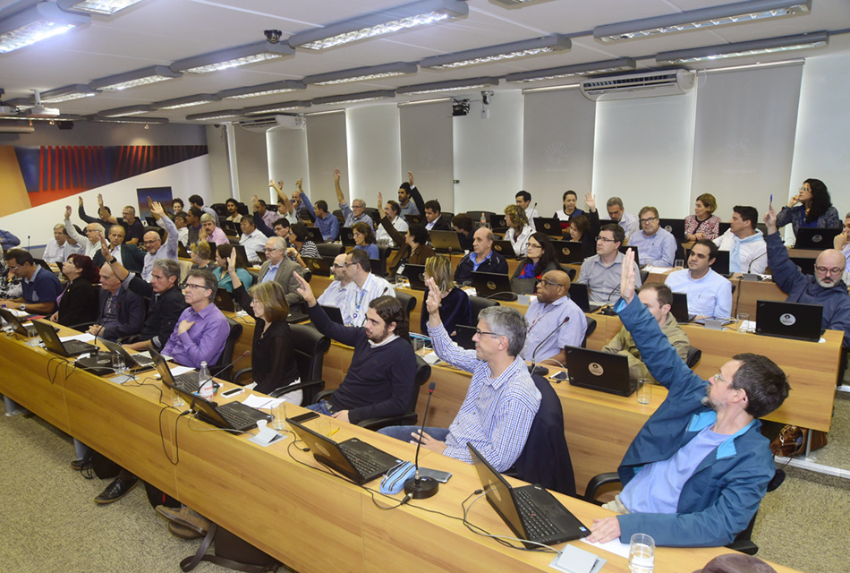 Em amplo auditório, em perspectiva, cerca de sessenta pessoas sentadas em cadeiras, uma ao lado da outros, formando cinco fileiras, a grande maioria com o braço erguido, durante votação. À frente deles, há extensas mesas retangulares de madeira sobre a qual há notebooks abertos, microfones de mesa, folhas de sulfite entre outros objetos. Imagem 5 de 6.