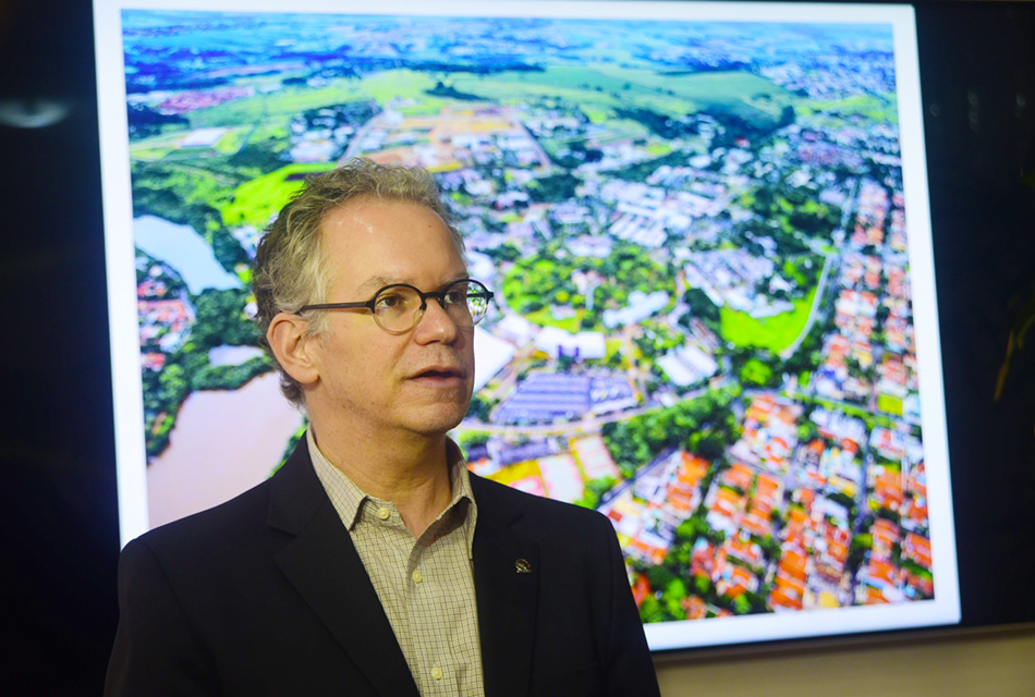 Homem, em pé, com os braços abaixados, de óculos e vestindo blazer preto sobre camisa social, sendo que às costas há uma foto colorida, com imagem aérea de área da Unicamp e do distrito de Barão Geraldo, onde está localizada. Imagem 6 de 6.