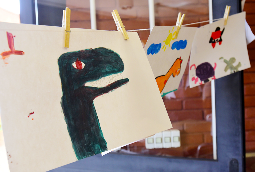 Em ambiente interno, imagem em perspectiva, parte de um varal com três ilustrações coloridas de dinossauros, feitas em folhas de cartolina, afixadas por pregadores. Em primeiro plano, há o pescoço e a cabeça de um dinossauro com a boca aberta, ilustrado em verde escuro. Imagem 9 de 10.