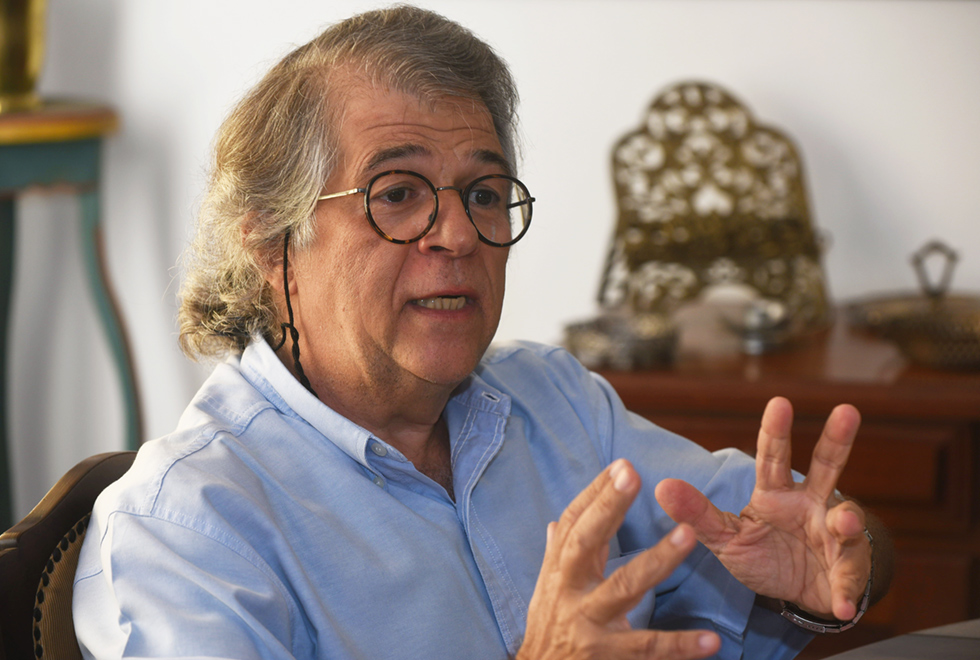 A galeria traz o sociólogo e professor da Unicamp Ricardo Antunes. Para acessá-la, tecle enter. Audiodescrição: Em sala de estar de residência, imagem em perspectiva e de busto, homem sentado em cadeira fala gesticulando com as mãos mantendo os braços levemente esticados para frente, com as mãos abertas, uma de frente para a outra. Ao fundo, há uma bancada de madeira sobre a qual estão um suporte para livros de metal e outras peças. Ele usa óculos e camisa social de mangas curtas em tom azul. Imagem 1 de 4.