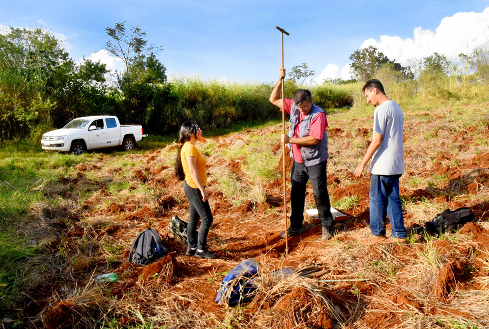 A galeria traz trabalho de campo de docente e alunos do Instituto de Geociências da Unicamp na Fazenda Argentina, em Campinas. Para acessá-la, tecle enter. Audiodescrição: Em campo aberto, área com vegetação rasteira capinada onde aparecem terra revolvida e mato seco, três pessoas em pé, sendo um homem ao centro, que perfura o solo com uma extensa vara de ferro, sendo observado por duas outras pessoas posicionadas junto a ele, à esquerda e à direita. Ao fundo, extensa área com mata. Imagem 1 de 5.