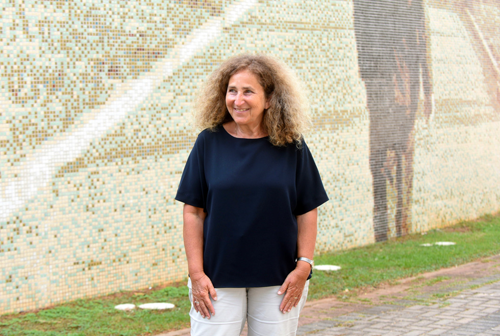 Num ambiente externo no fundo parte de um trabalho de mosaico onde aparece um caminho e uma pessoa sem a cabeça de costa veste terno escuro. Na frente do painel de mosaico um canteiro gramado seguindo um chão com blocos de cimento. Uma pessoa aparecendo de meio corpo veste camiseta preta com calça jeans claro. No seu braço esquerdo ostenta um relógio.