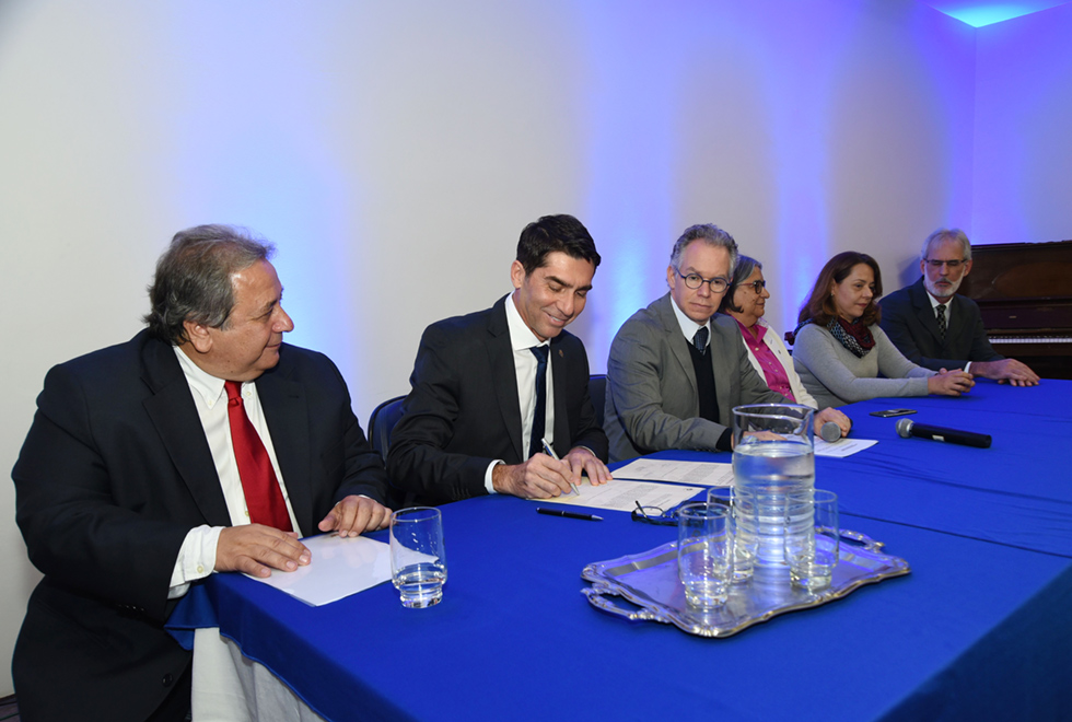 A galeria mostra a cerimônia de posse do professor José Roberto Ribeiro como novo diretor do Colégio Técnico de Limeira (Cotil). para acessá-la, tecle enter. Audiodescrição: Ambiente interno, no fundo parede branca vários pontos de luz verde claro, parte um piano. A frente mesa retangular com pano branco e azul. Seis pessoas sentadas a mesa, esquerda para direita, a segunda pessoa escreve numa folha de papel, sendo observado pelos demais. Bandeja com jarra de água, copos, óculos, caneta e microfone na mesa.