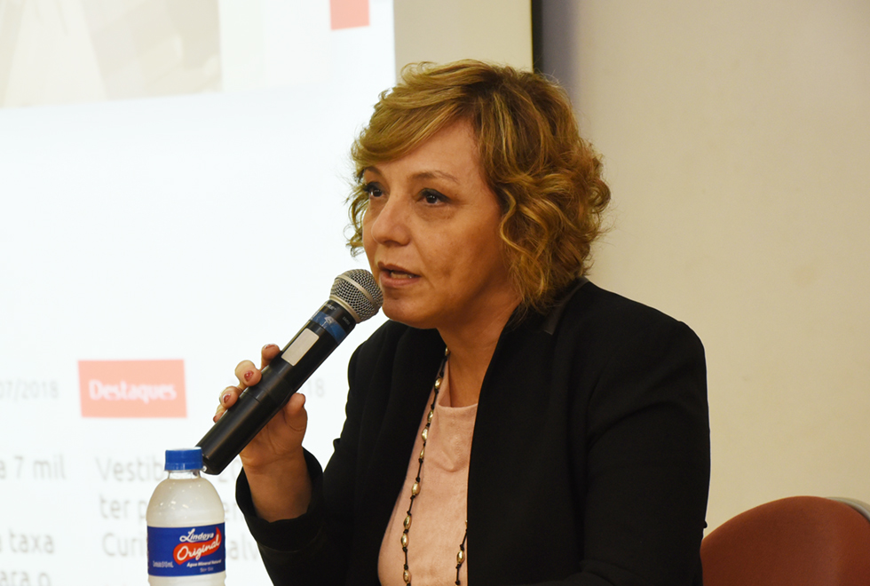 Em auditório, imagem em perspectiva e de busto, mulher sentada em cadeira fala em microfone sem fio, segurando-o com a mão direita, enquanto mantém o braço esquerdo baixado. Há uma garrafa pet pequena de água apoiada sobre suposta mesa. Ao fundo, afixada na parede, aparece parte de uma tela de projeção de data show. Imagem 3 de 8.