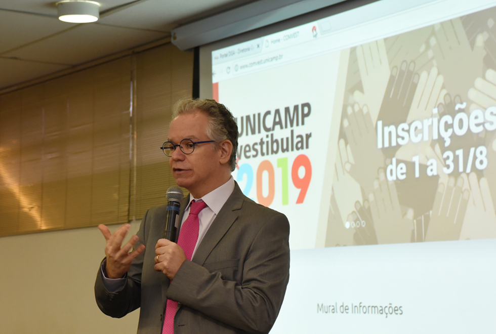 A galeria traz o lançamento do Vestibular Unicamp 2019 e do novo curso de Engenharia do Transporte. Para acessá-la, tecle enter. Audiodescrição: Em auditório, imagem em perfil e plano médio, homem em pé, ao centro, fala em microfone sem fio segurando-o com a mão esquerda, enquanto gesticula com a direita, mantendo o braço dobrado e projetado para a esquerda. Às costas dele, há uma tela de projeção de data show onde se lê Unicamp Vestibular 2019. Ele usa óculos e veste terno marrom e gravata. Imagem 1 de 8.