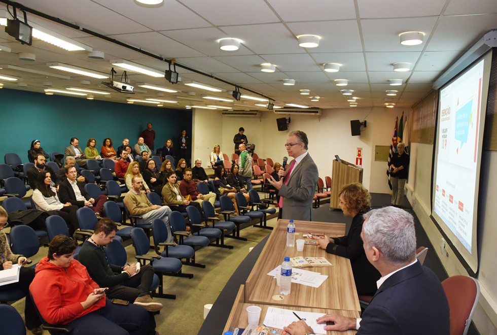 No palco de um auditório, imagem em perfil e de corpo inteiro, homem em pé fala em microfone sem fio, sendo que à esquerda dele há duas pessoas sentadas em cadeiras, uma ao lado da outra, tendo à frente delas uma mesa/balcão de madeira. Ambos mantém braços apoiados sobre a mesa. Ao fundo, às costas deles, à direita na imagem, há uma tela de projeção de datashow. À frente deles, o público do auditório, cerca de trinta pessoas, que se encontra sentado em cadeiras dispostas uma ao lado da outra. Imagem 5 de 8.