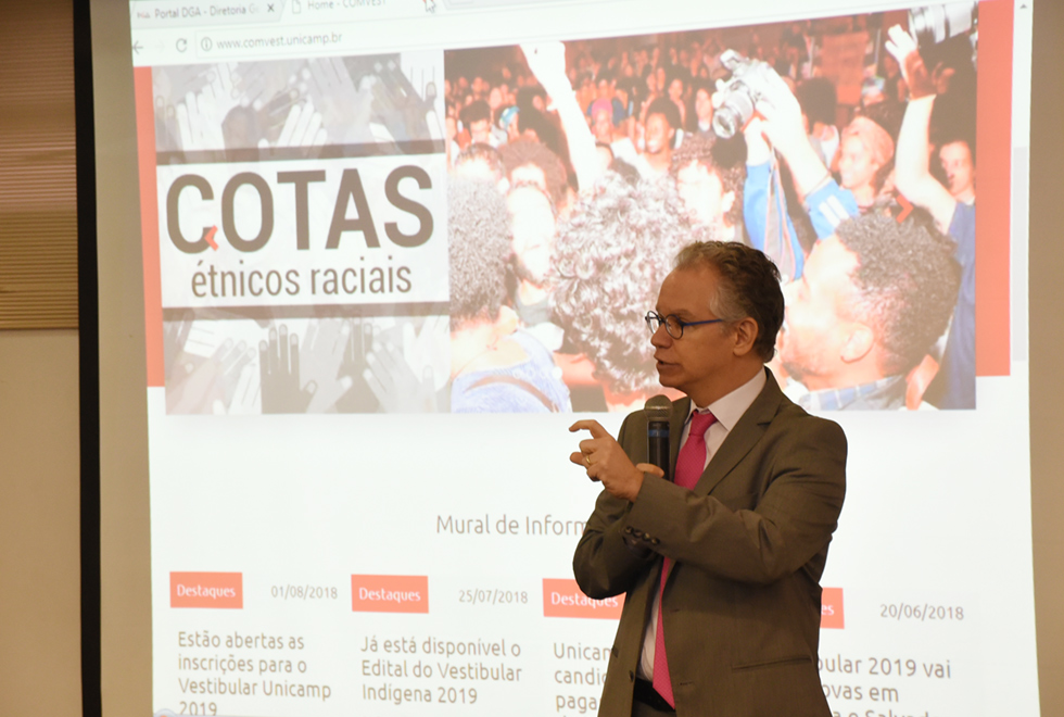 Em auditório, imagem em perfil e plano médio, homem em pé, à direita na imagem, fala em microfone sem fio segurando-o com a mão direita, enquanto gesticula com a esquerda, mantendo o braço dobrado e projetado para frente, e o dedo indicado curvado, em forma da letra c. Às costas dele, há uma tela de projeção de data show onde se lê cotas étnicos raciais. Ele usa óculos e veste terno marrom e gravata. Imagem 6 de 8.