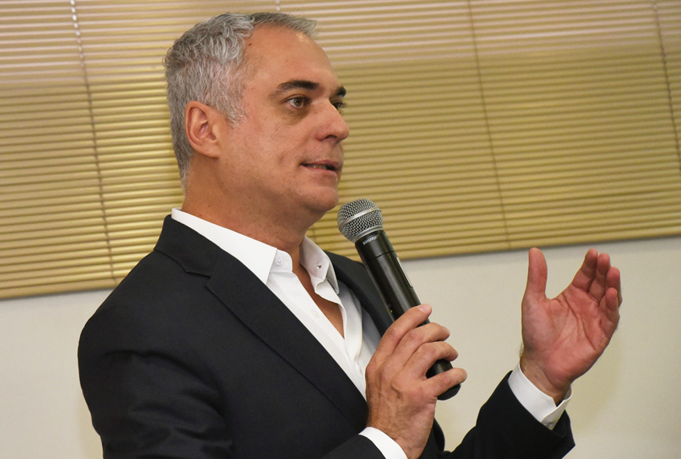 Em área interna, imagem em perspetiva e de busto, homem em pé, à esquerda na imagem, fala em microfone sem fio, segurando-o com a mão direita, enquanto gesticula com o braço esquerdo, mantendo o braço dobrado e levemente projetado para frente, com a palma da mão aberta. Às costas dele há persianas horizontais. Ele veste blazer preto sobre camisa social branca. Imagem 7 de 8.
