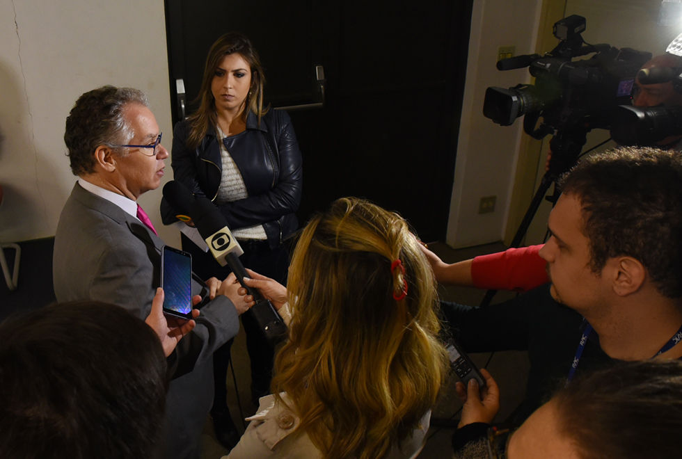 Em área interna, imagem em perfil e plano médio, homem em pé, À esquerda na imagem, concede entrevista coletiva, sendo que ele está cercado por repórteres que seguram nas mãos equipamentos como microfone, celular e gravador digital, mantendo o braço esticado para frente, na direção do entrevistado. O ambiente possui pouca luminosidade. O entrevistado usa óculos, terno cinza e gravata. Imagem 8 de 8.