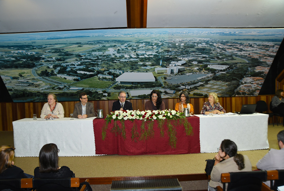 Ambiente interno, no fundo parede azul escuro, branco com madeira nela painel com fotografia aérea da Unicamp. Primeiro plano mesa retangular coberta por pano branco outro menor vermelho tendo em cima arranjo flores brancas, vermelhas e ramos verdes. Sentados a mesa encontram seis pessoas, terceira pessoa, esquerda para direita, usando óculos segura com a mão direita microfone perto da boca. Os demais prestam atenção. Do lado mesa pessoa sentada em cadeira. Frente da mesa aparece quatro pessoas em assentos.