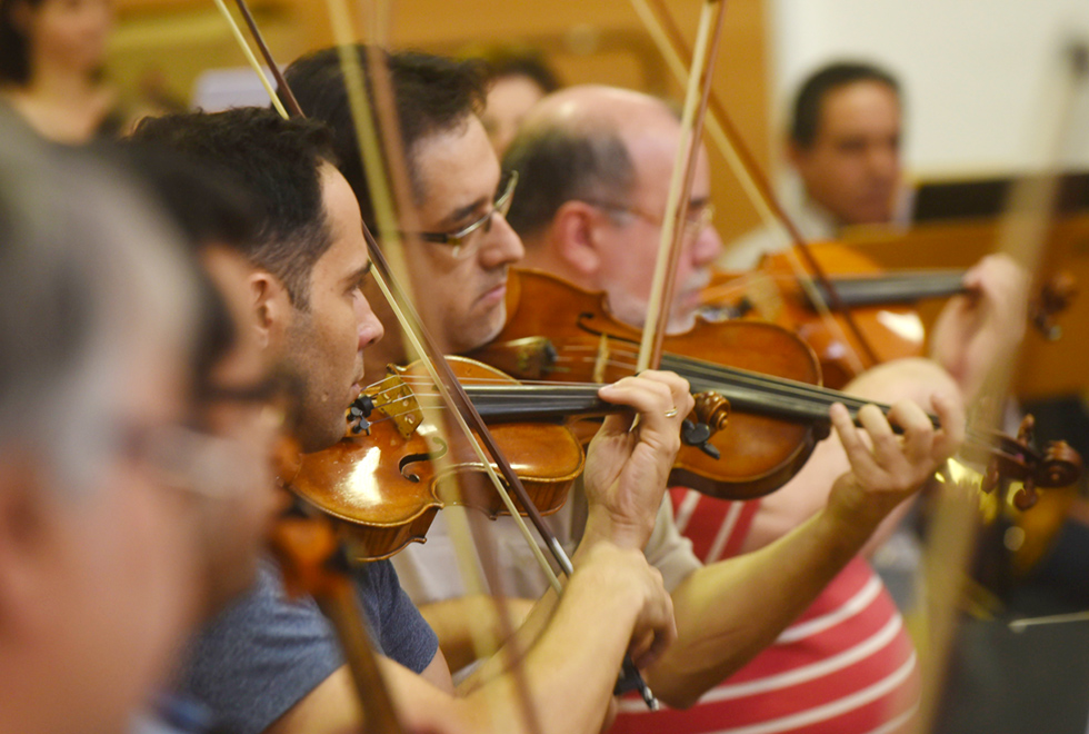 Em área interna, imagem close-up e em perspectiva, três homens tocam violino, dispostos um ao lado do outro, sendo que estão posicionados da esquerda para a direita, com olhares voltados para a direita. Imagem 3 de 9.