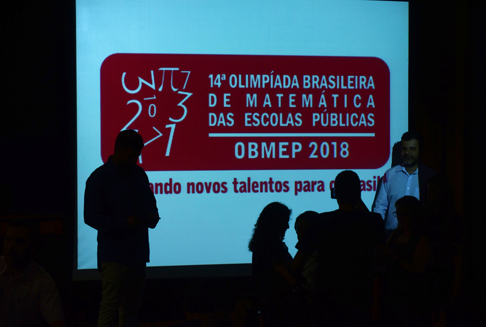 Num ambiente interno, o fundo esta escuro com uma tela branca, onde destaca, 14ª olimpíada brasileira de matemática das escolas públicas - OBMEP 2018. De frente para tela, partes de pessoas, que aparecem todas escuras. Do lado direito, da tela, parte de uma pessoa, vestindo uma camisa branca. Imagem 6 de 13